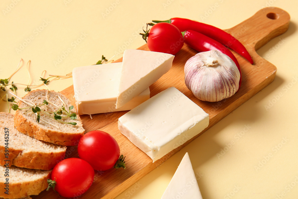 Board with processed cheese, bread and vegetables on pale yellow background