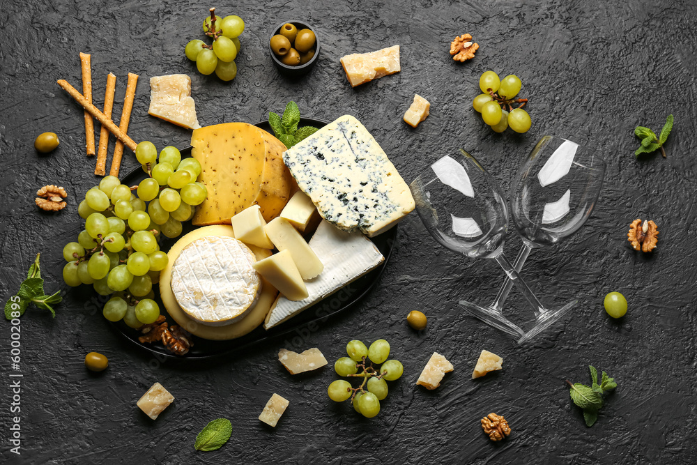 Tray with different types of tasty cheese, grapes and wine glasses on dark background