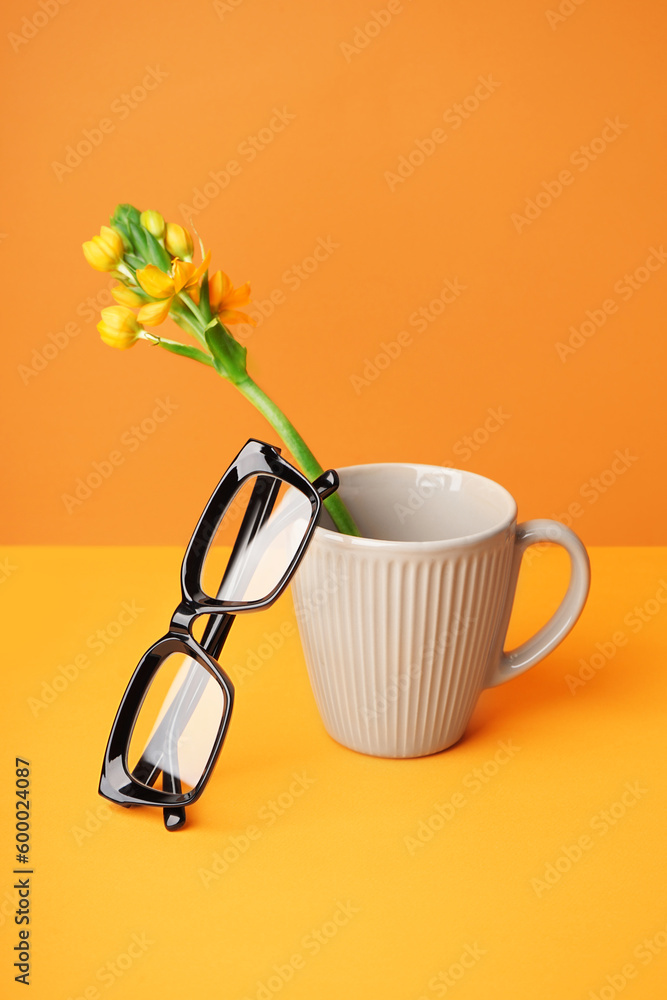 Stylish eyeglasses and cup with flower on yellow table near wall