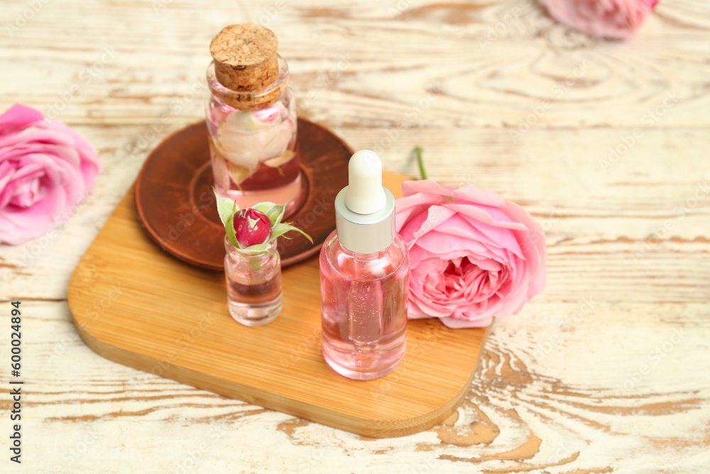 Bottles of cosmetic oil with rose extract and flowers on light wooden table