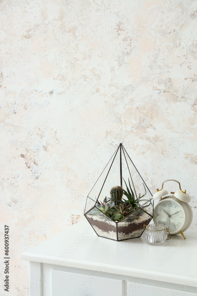 Florarium with alarm clock and candle on table near light wall