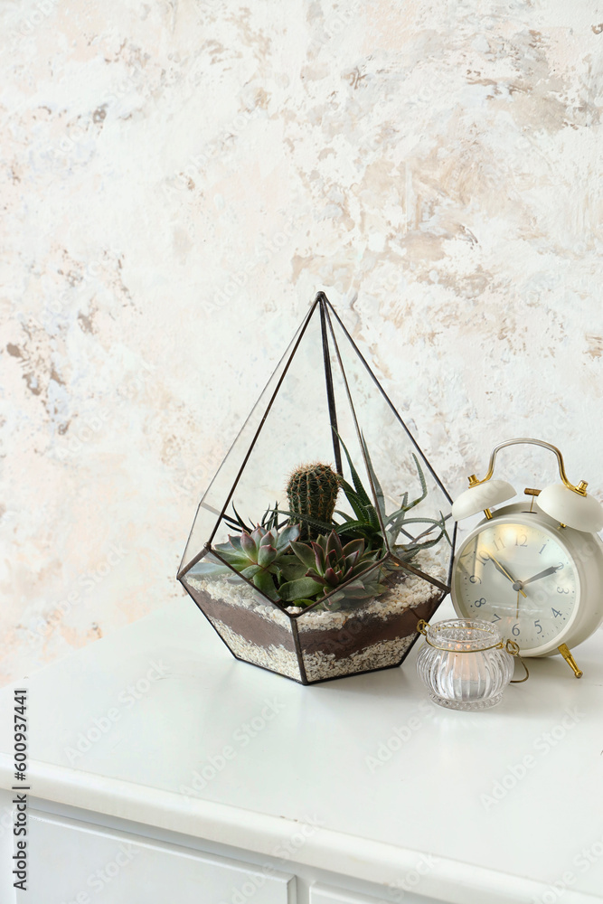 Florarium with alarm clock and candle on table near light wall, closeup