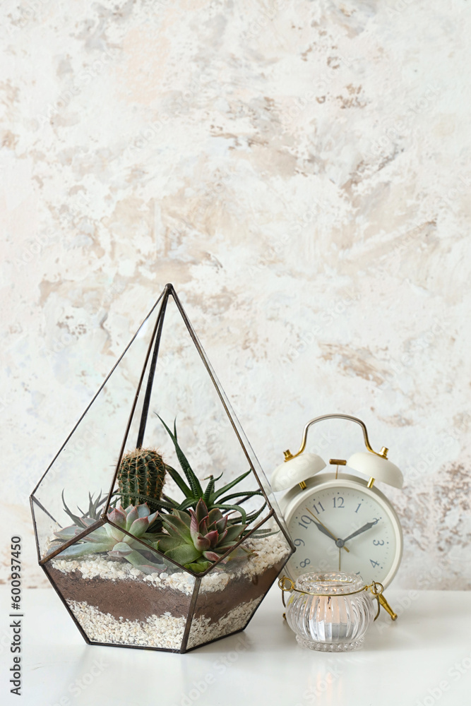 Florarium with alarm clock and candle on table near light wall