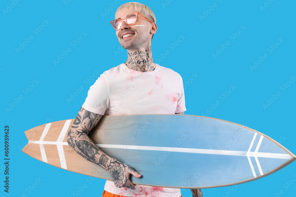 Young tattooed man with surfboard on light blue background