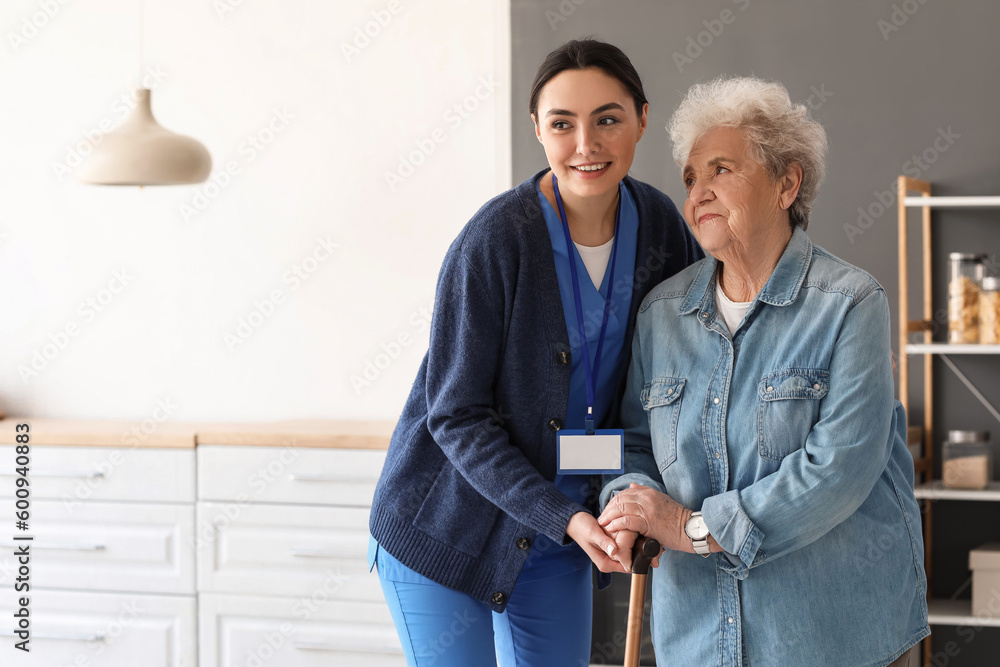 Young caregiver helping senior woman with walking stick in kitchen