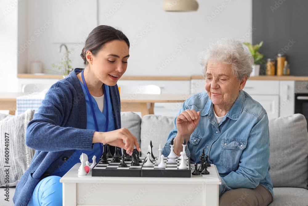 Young caregiver with senior woman playing chess at home