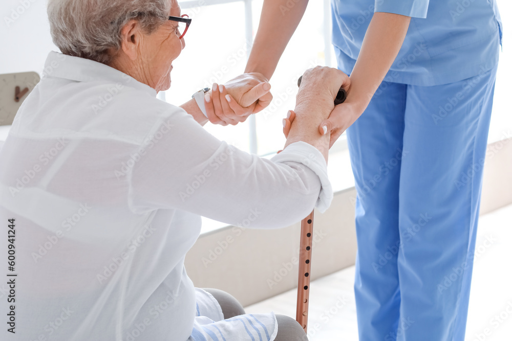 Young caregiver helping senior woman with stick to stand up in bedroom, closeup