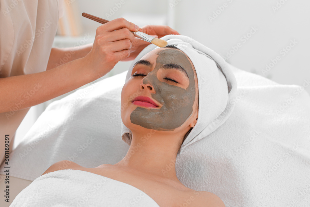 Cosmetologist applying clay mask onto young womans face in salon, closeup