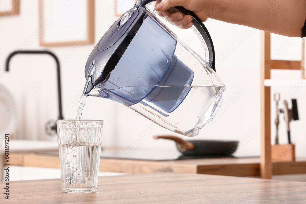 Woman pouring pure water from filter jug into glass on table in kitchen