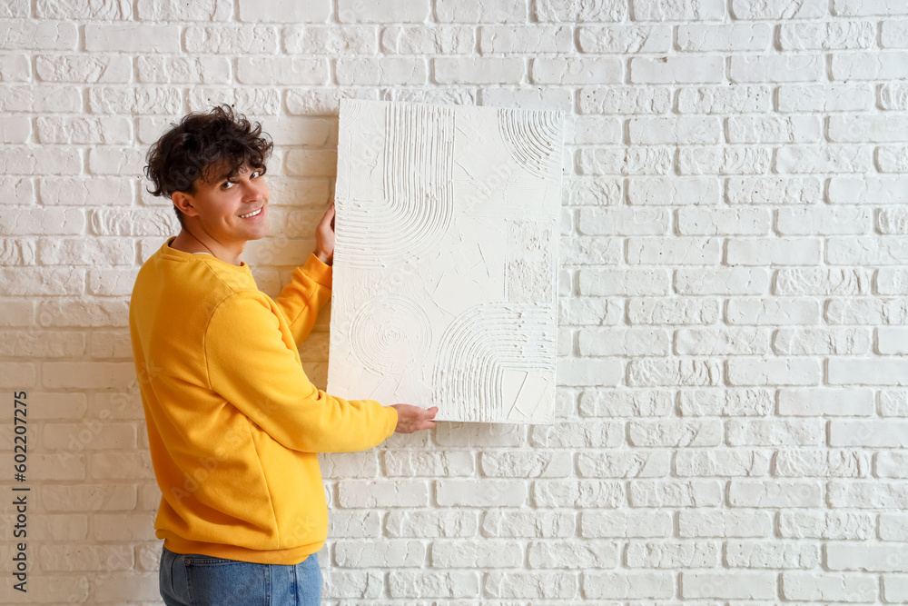 Young man hanging painting on white brick wall