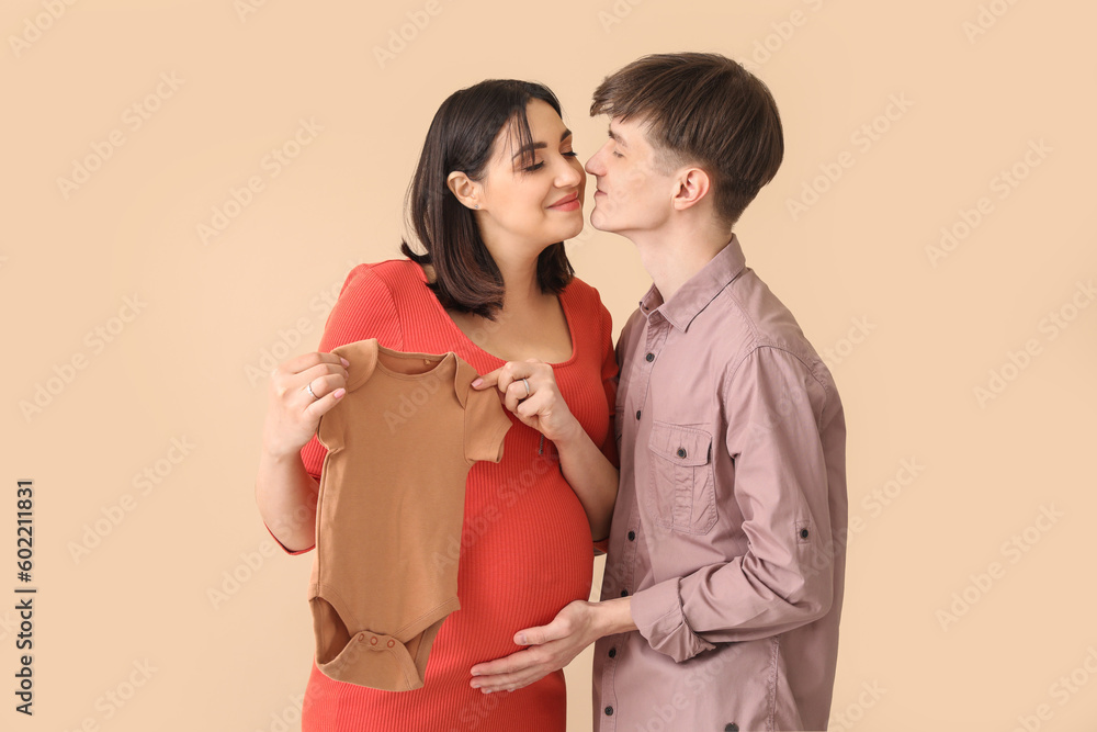 Young pregnant couple with baby bodysuit on beige background