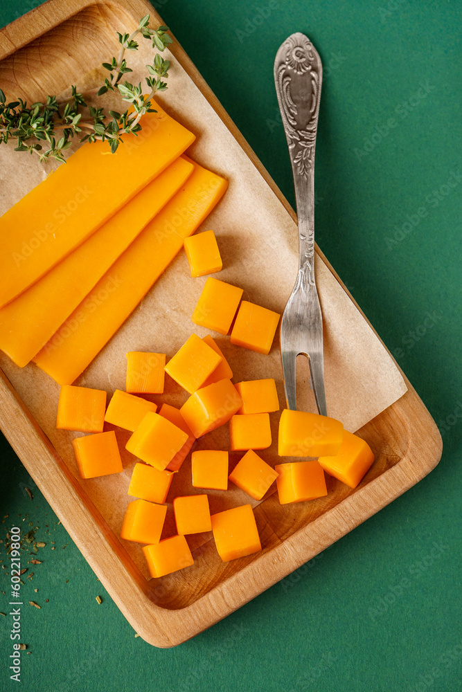 Board with pieces of tasty cheddar cheese and herbs on green background