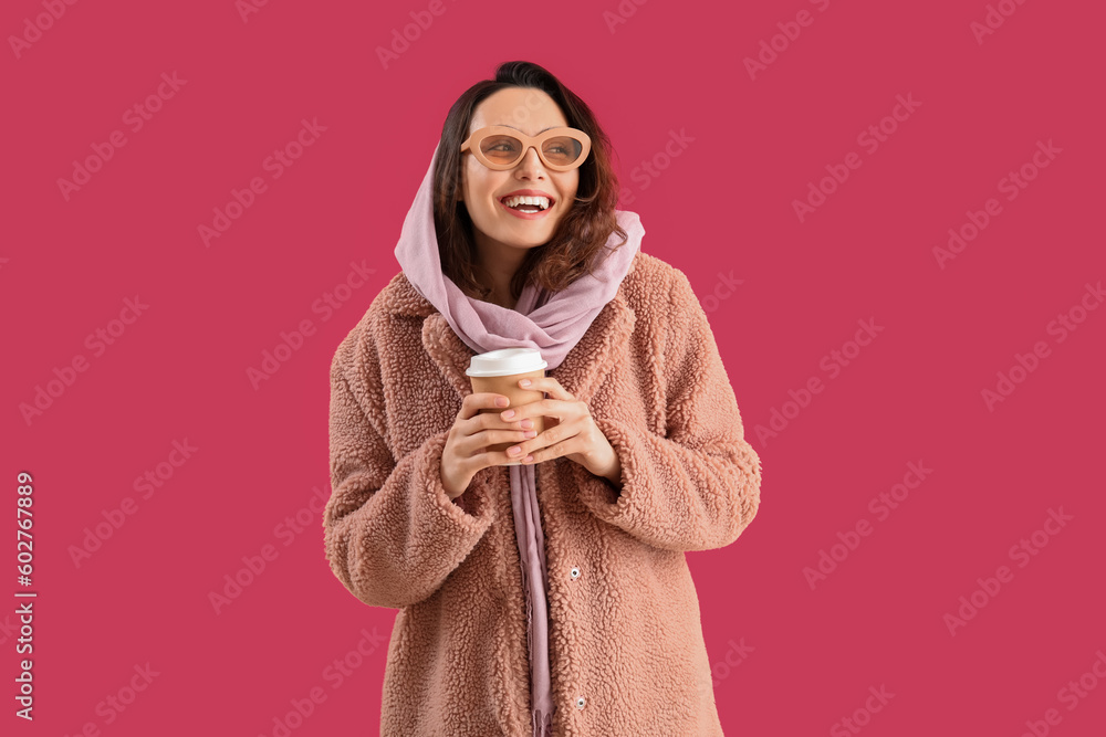 Stylish young woman in teddy coat with cup of coffee on pink background