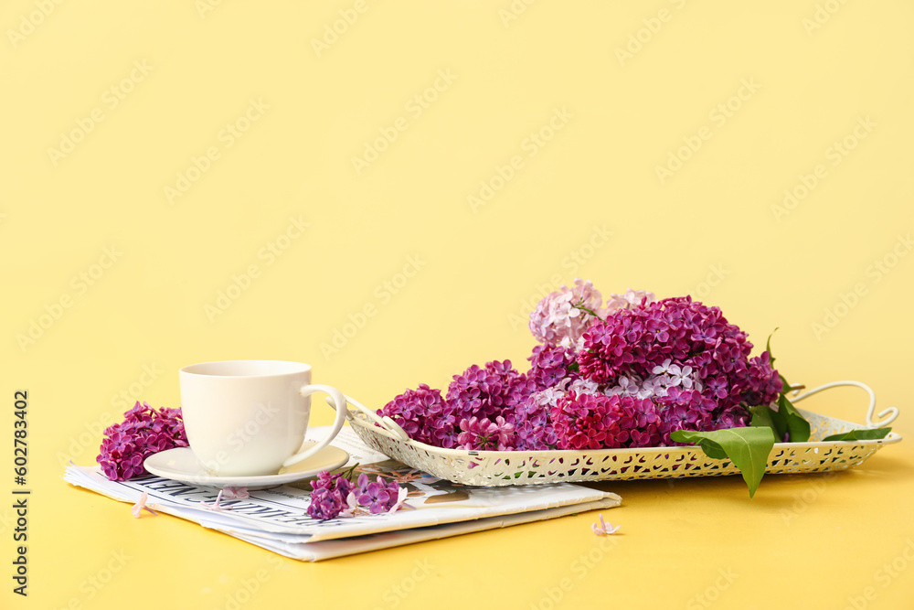 Cup of tea with beautiful fragrant lilac flowers and magazine on pale yellow background