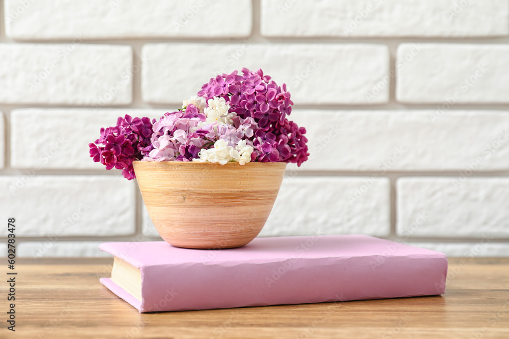 Wooden vase of beautiful fragrant lilac flowers and book on wooden table near white brick wall