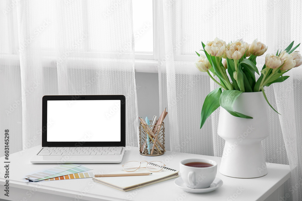 Comfortable workplace with modern laptop, cup of tea and beautiful tulip flowers near window