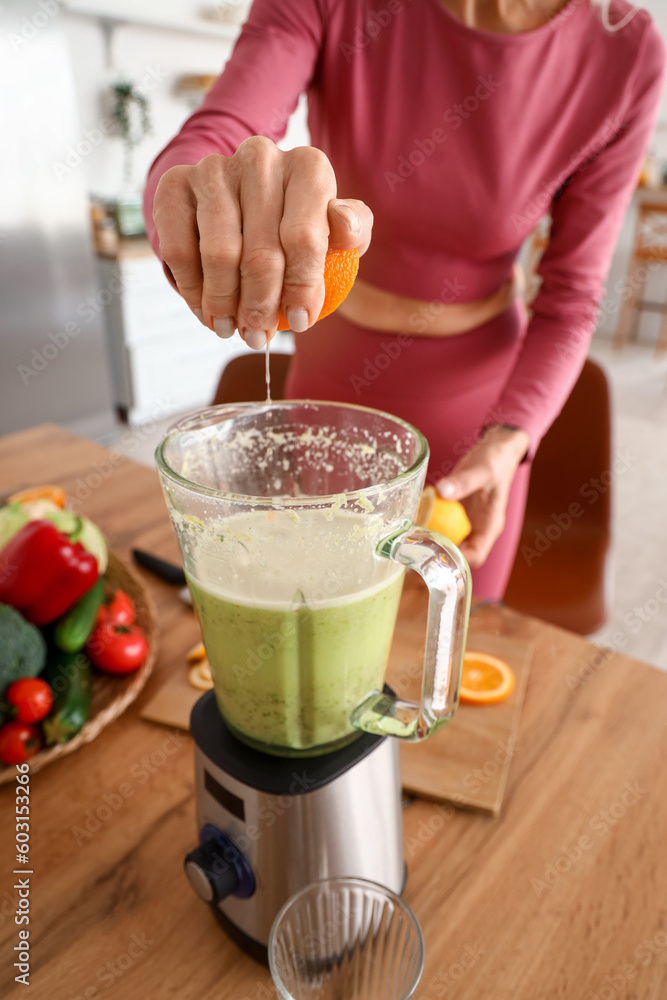 Sporty mature woman squeezing orange juice into blender with healthy smoothie in kitchen, closeup