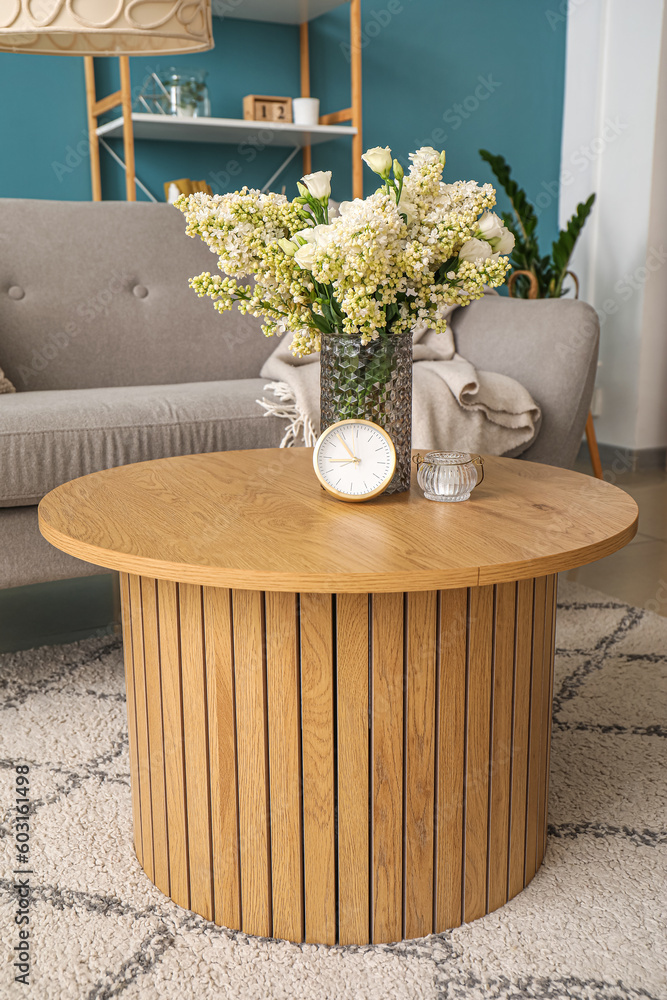 Vase with blooming lilac flowers and alarm clock on wooden coffee table in living room