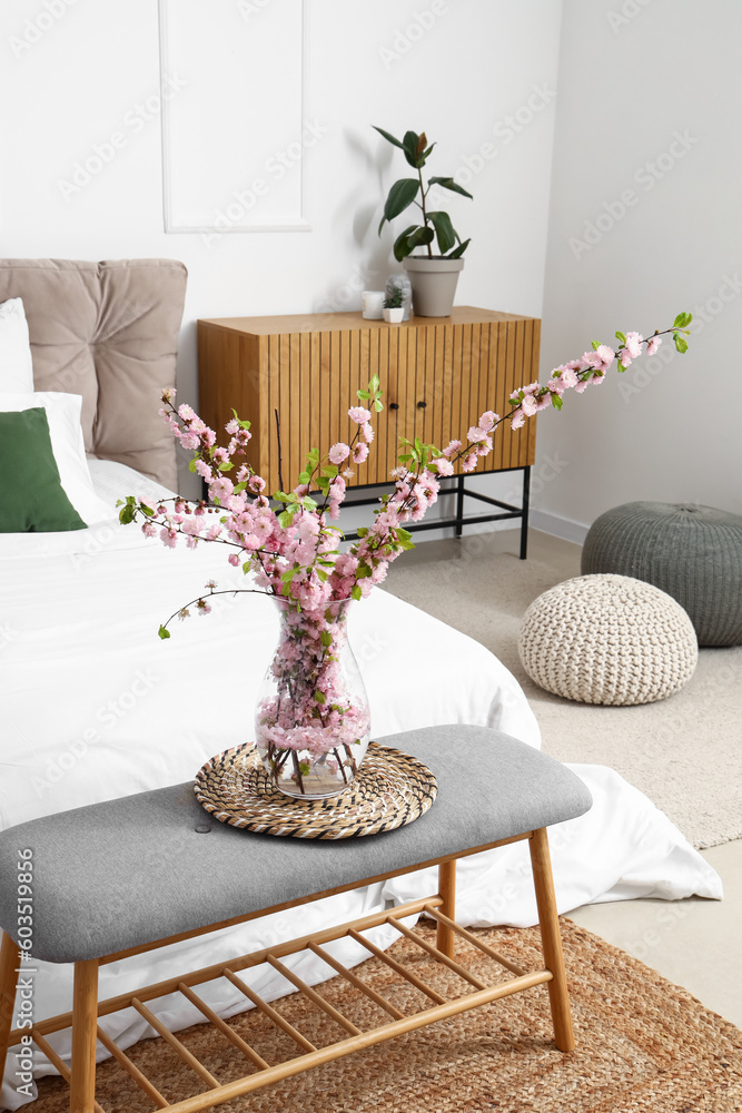 Vase with blooming sakura branches on bedside bench in interior of bedroom