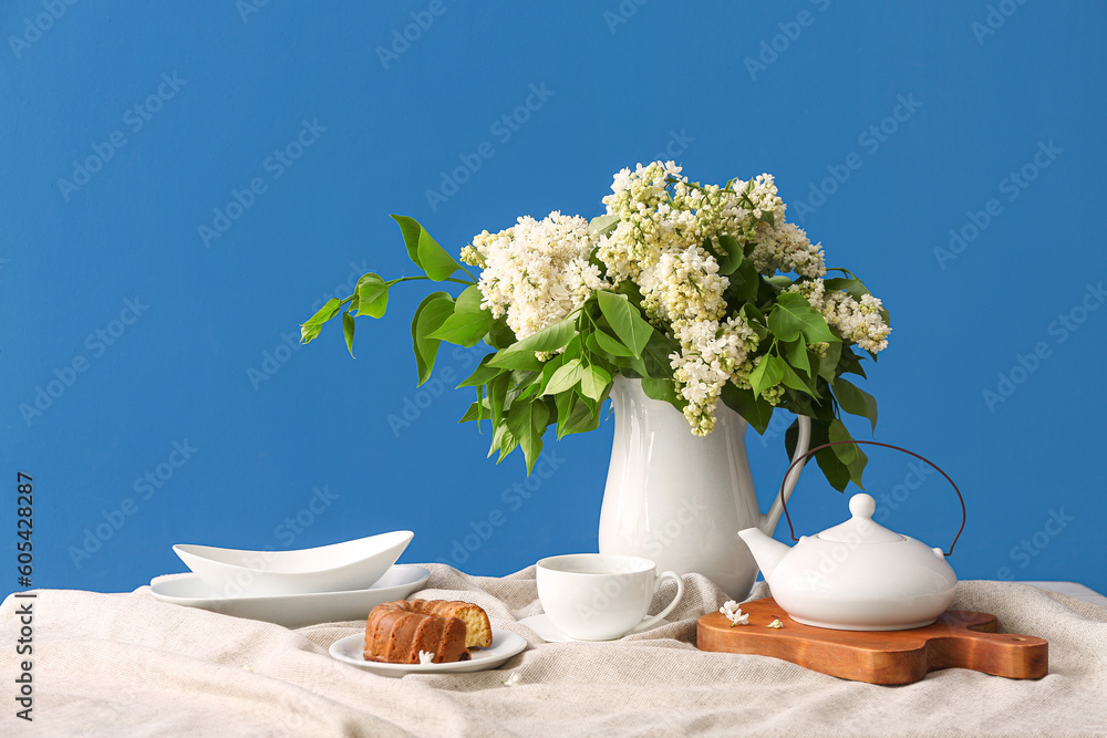 Composition with vase of white lilacs, cup of coffee and cake on table near blue wall