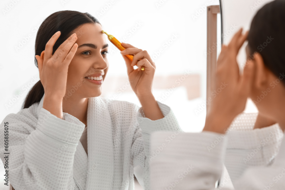 Young woman with facial massage tool near mirror in bathroom