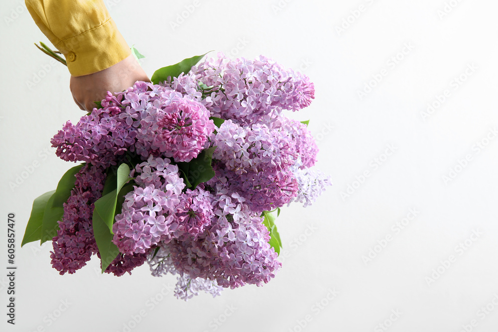 Female hand holding bouquet of beautiful lilac flowers near white wall