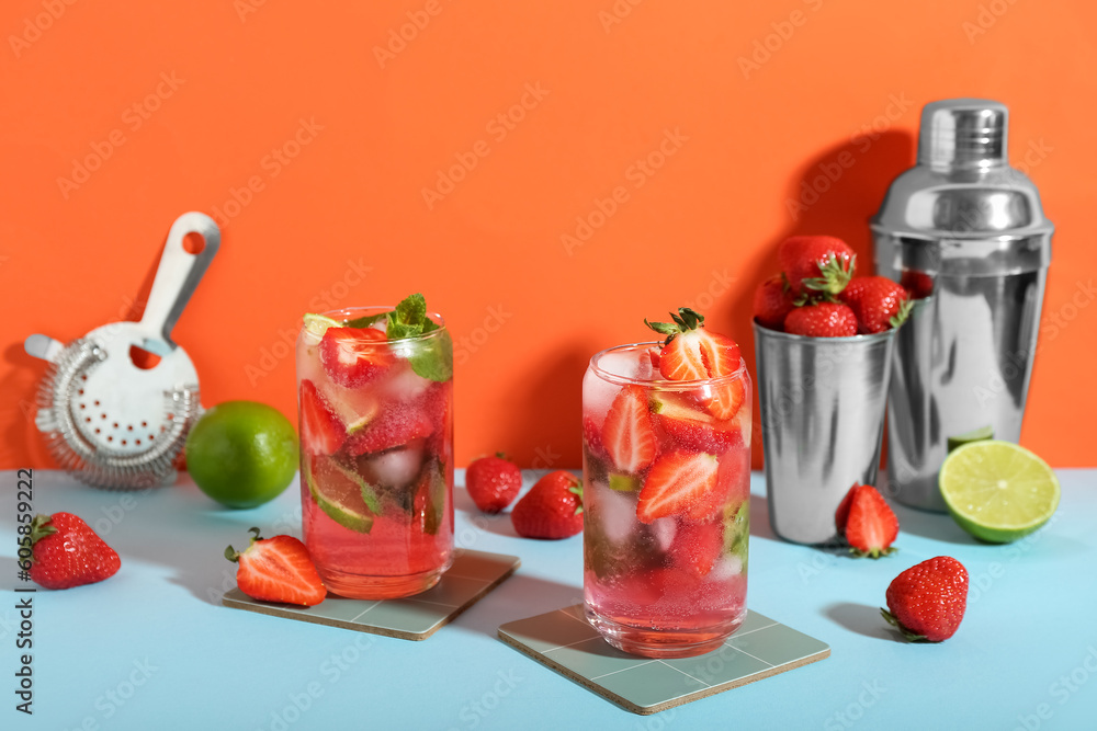 Glasses of fresh strawberry mojito with shakers, strainer and ingredients on blue table near orange 