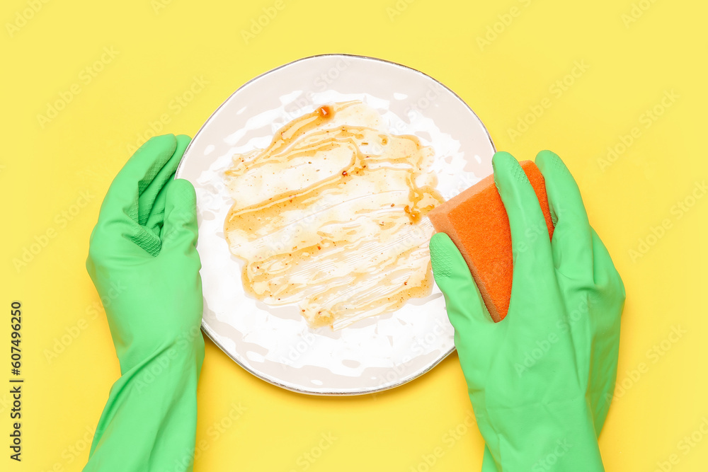 Female hands in rubber gloves washing dirty plate on yellow background