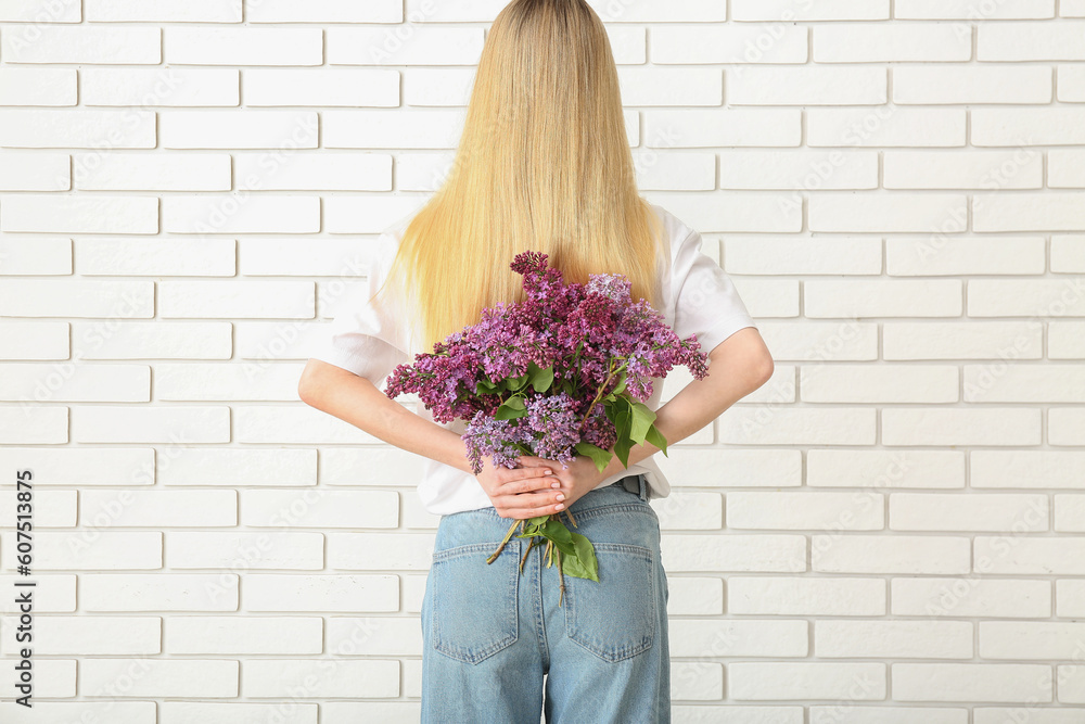 Woman holding bouquet of lilacs behind her back near white brick wall