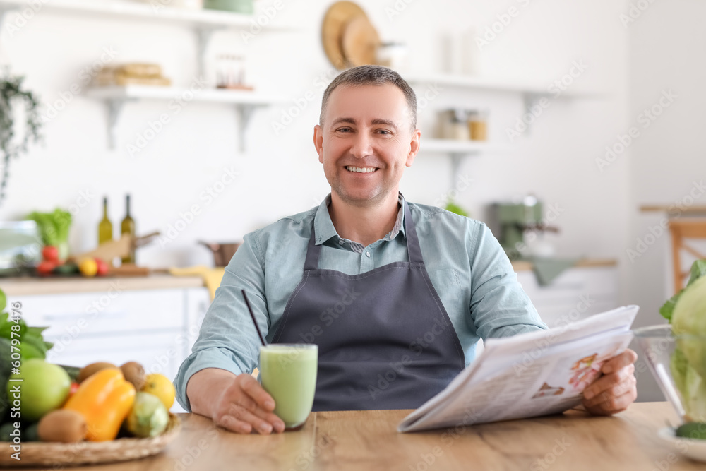 Mature man with glass of fresh vegetable smoothie reading newspaper in kitchen