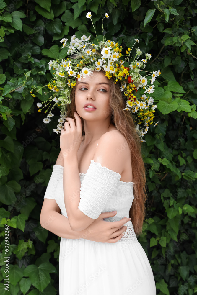 Beautiful young woman in flower wreath near green ivy outdoors. Summer solstice