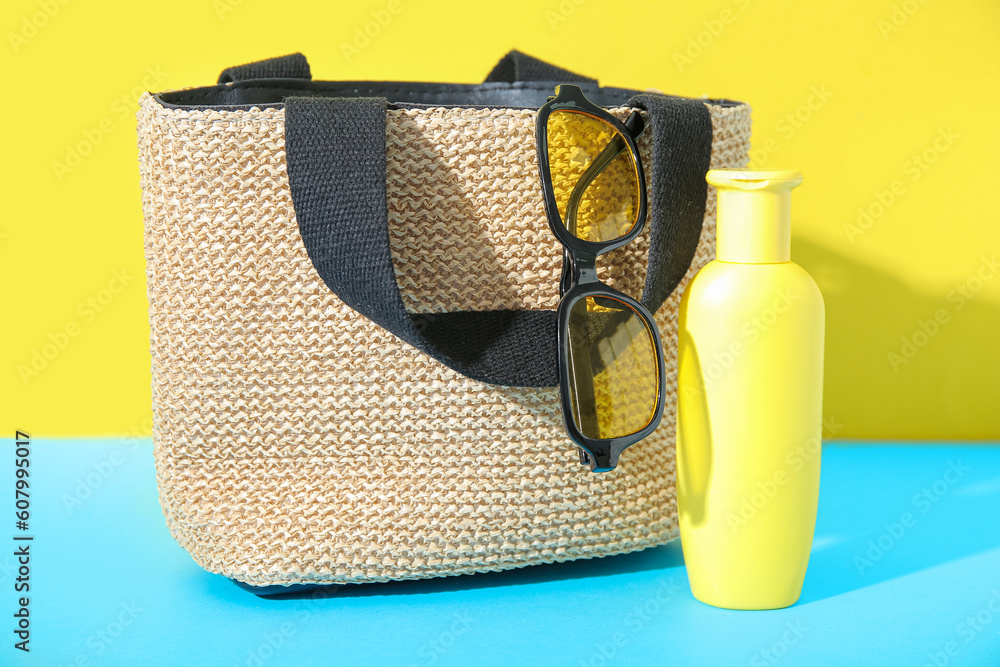 Bottle of sunscreen cream with sunglasses and wicker bag on blue table near yellow wall