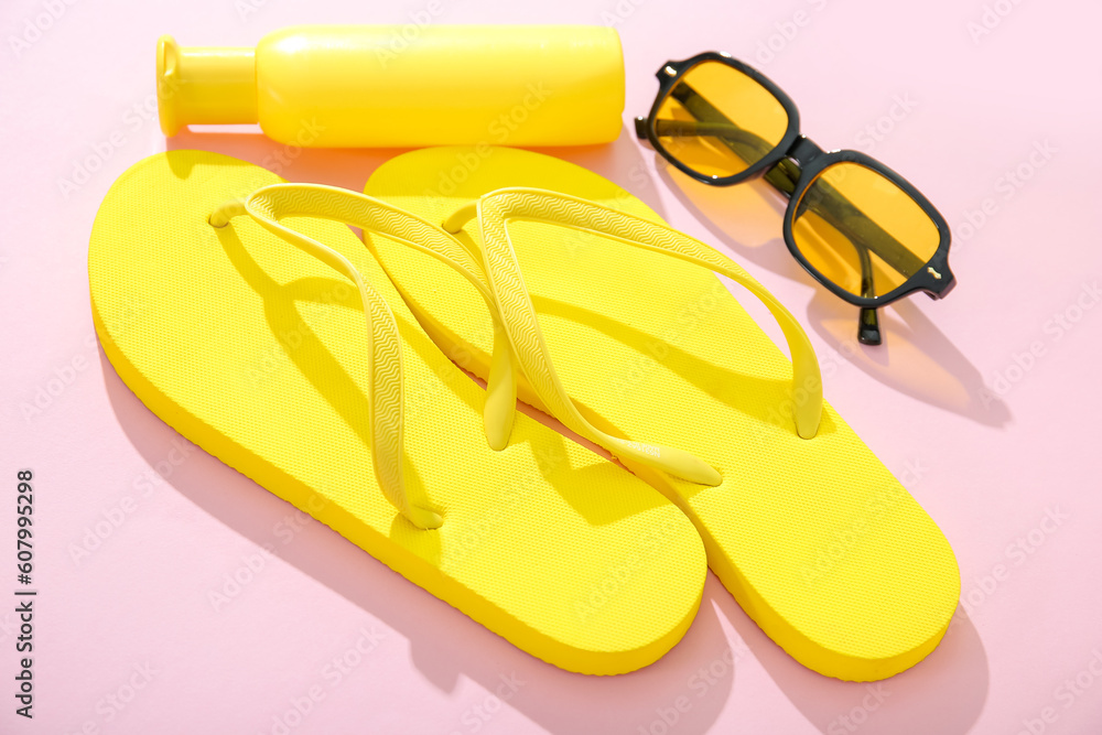 Bottle of sunscreen cream with sunglasses and flip flops on pink background