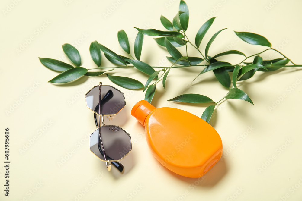 Bottle of sunscreen cream with sunglasses and tree branch on pale yellow background