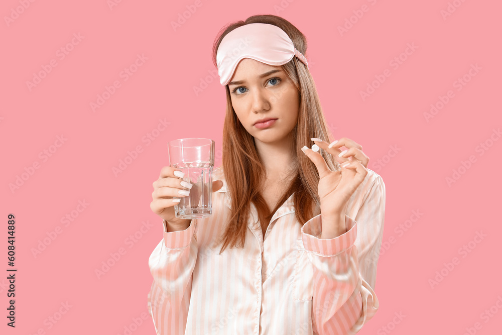 Upset young woman in pajamas with pill and glass of water on pink background