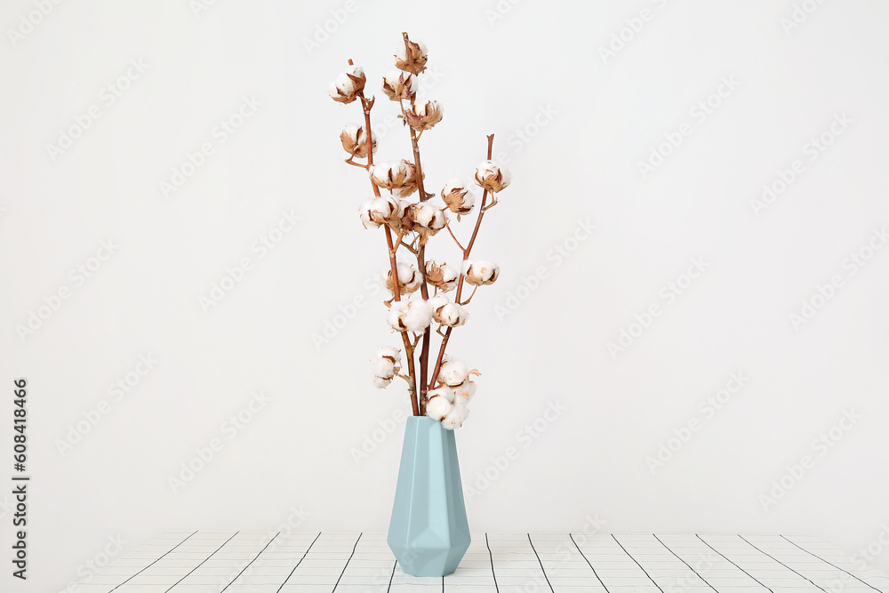 Cotton sprigs in vase on white tile table near wall