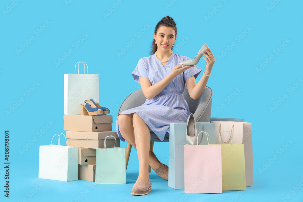 Young woman with shoe boxes and shopping bags sitting in armchair on blue background