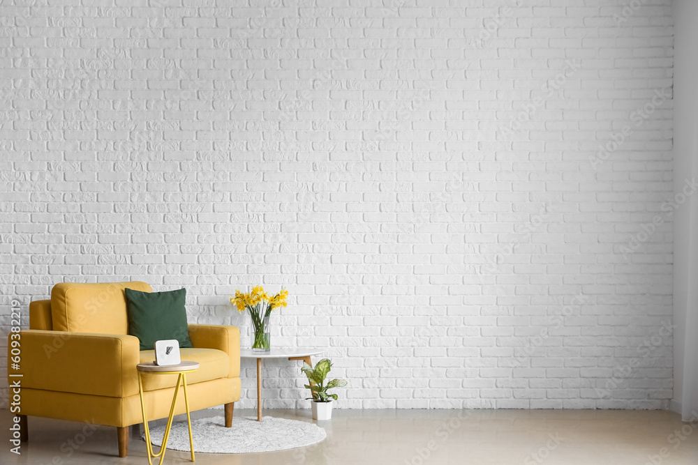 Cozy yellow armchair and vase with blooming narcissus flowers on coffee table near white brick wall