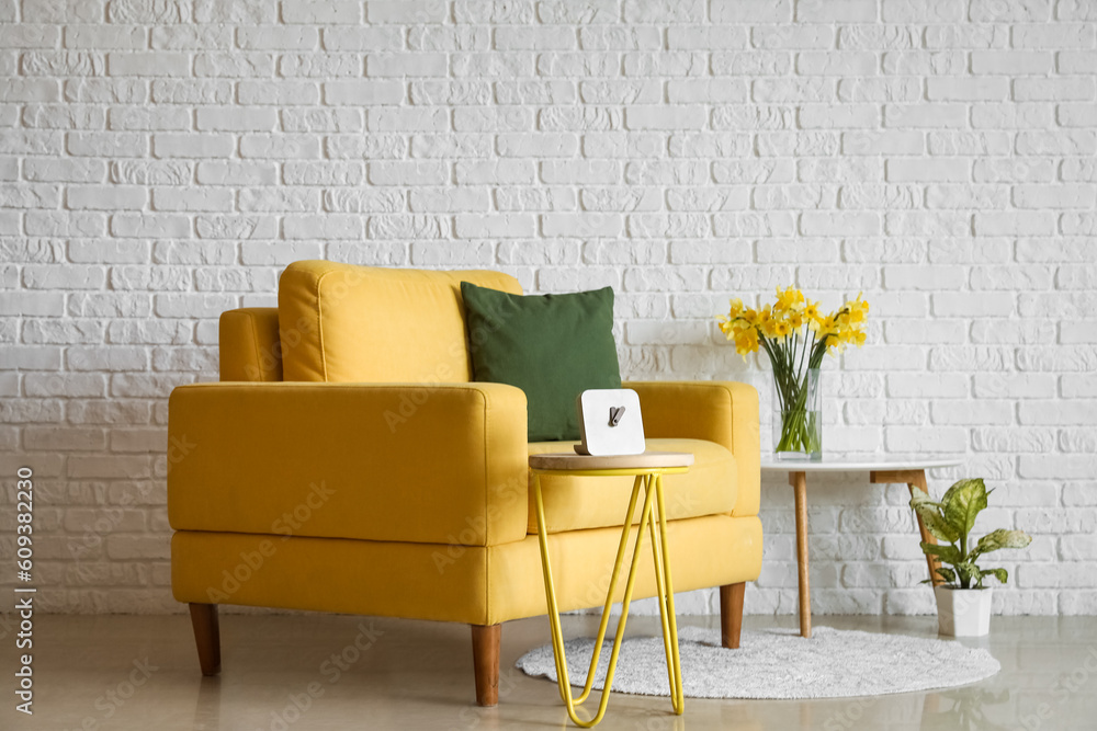 Cozy yellow armchair and vase with blooming narcissus flowers on coffee table near white brick wall