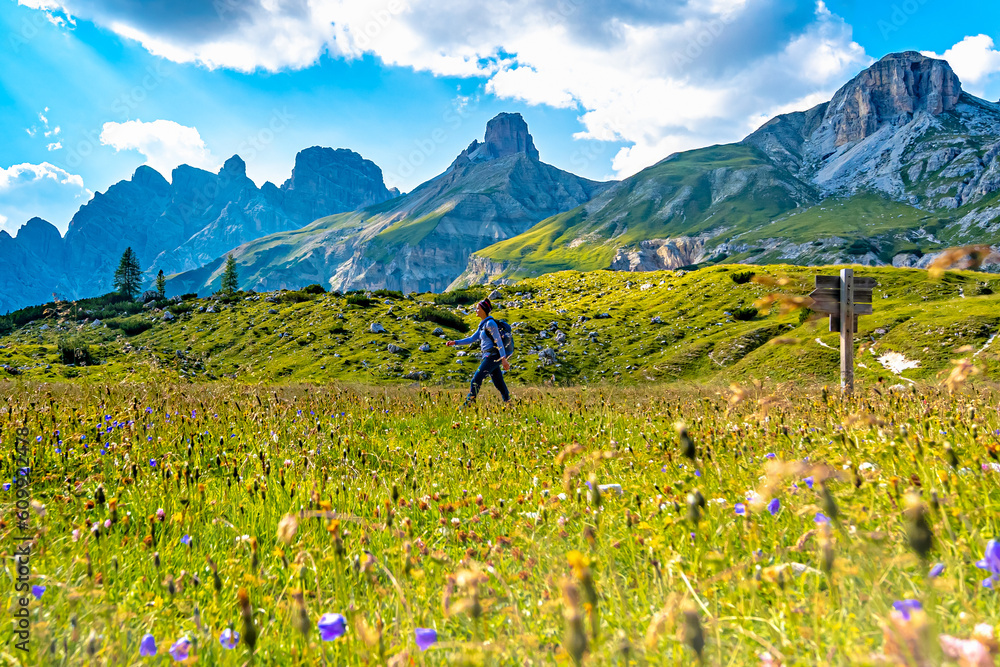 Woman walking along scenic Dolomites high trail with meadow in the foreground in the afternoon. Tre 