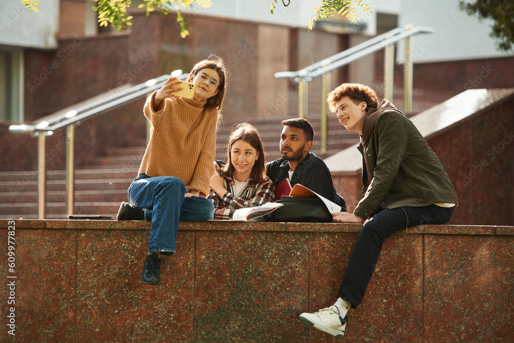 Woman is making selfie. Four young students in casual clothes are together outdoors