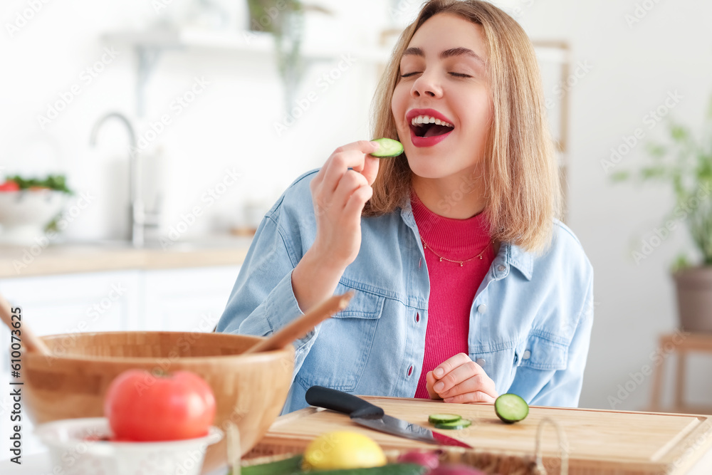 Young woman eating cucumber slice while making salad in kitchen