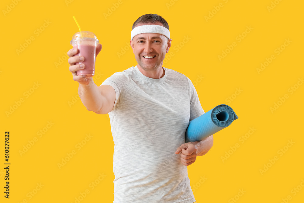 Sporty mature man with glass of fruit smoothie and fitness mat on yellow background