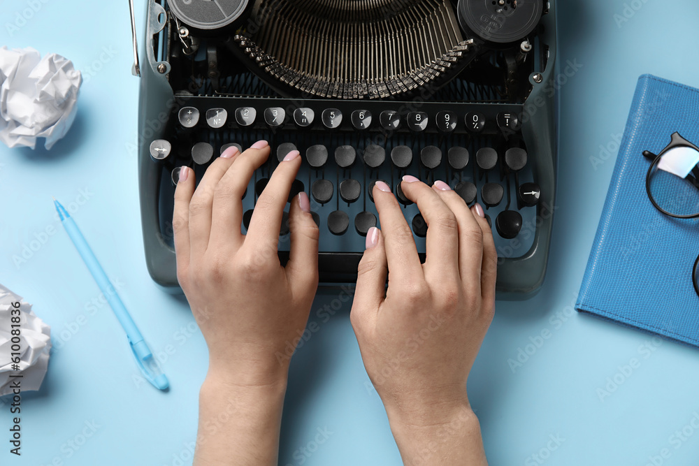 Woman typing on typewriter with crumpled paper, notebook and eyeglasses on light blue background