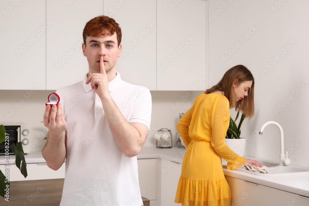 Young man with engagement ring showing silence gesture before proposing to his girlfriend in kitchen