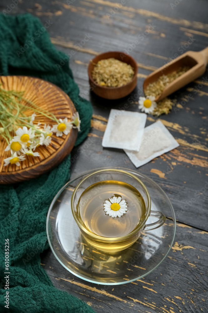 Cup of hot tea and bowl with dried chamomile on black wooden background