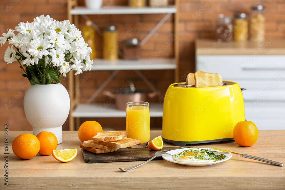 Modern toaster, glass of juice, oranges and fried eggs on table in kitchen