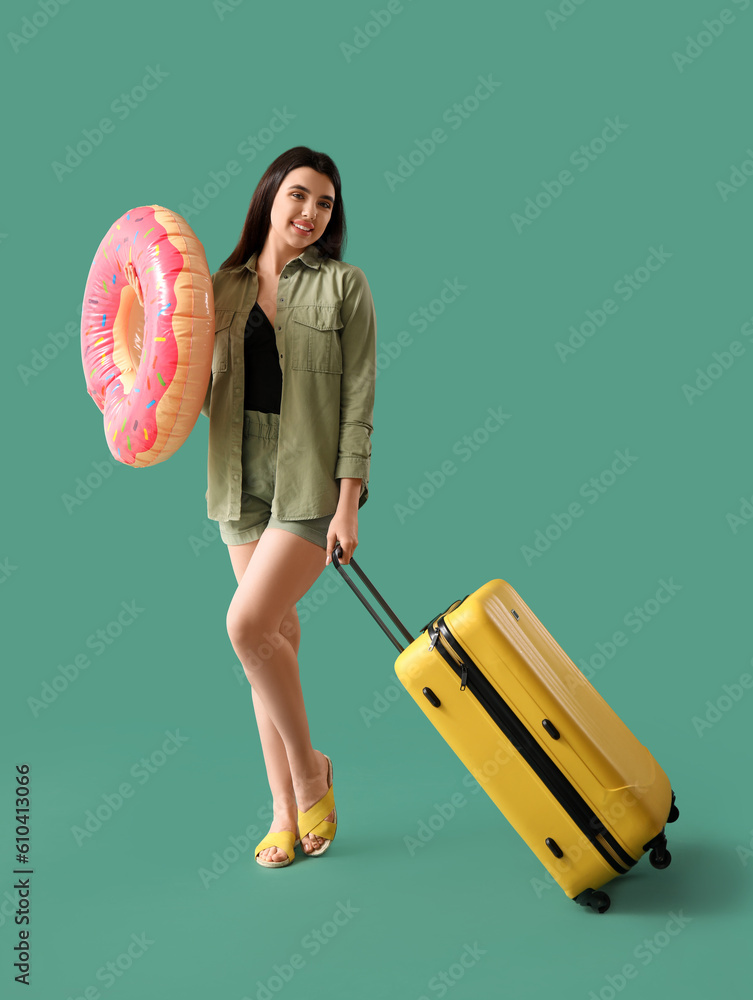 Young woman with inflatable ring and suitcase on green background