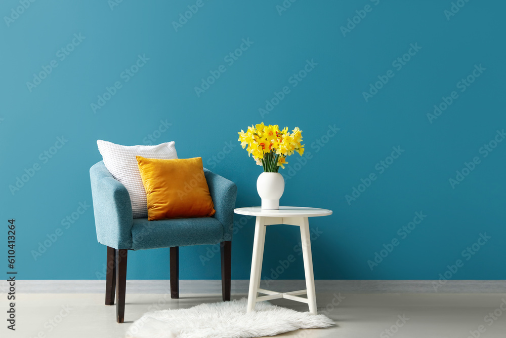 Cozy armchair with cushions and blooming narcissus flowers on coffee table near blue wall