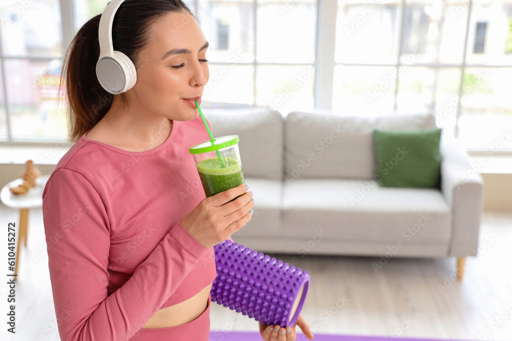 Sporty young woman in headphones drinking vegetable juice at home